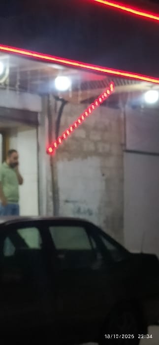 The image depicts a nighttime urban scene in Al-Kiswa, Syria, dated 18/10/2025 at 22:34. A dark-colored vehicle, possibly a sedan, occupies the lower foreground, presenting its roofline and windows from the front passenger side. In the midground, to the left, a bearded male wearing a light green shirt and blue jeans stands in an open doorway or recessed area, holding a dark object to his right ear, indicating a phone call. The background consists of a building facade with exposed brickwork partially painted light. Overhead, two bright circular white lights illuminate the area. Prominently, red neon strip lighting is affixed to the building: two parallel horizontal strips along the top, and a diagonal strip composed of individual red light dots extending downwards from the right. The overall environment is dimly lit by artificial sources.