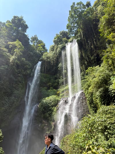 The image captures a majestic waterfall in the lush, green surroundings of Sawan, Indonesia. The waterfall, appearing as multiple cascading streams of white water, plunges down a rocky, vegetation-covered cliff face. The scene is framed by dense foliage of trees and plants, with sunlight filtering through the leaves, illuminating the mist rising from the base of the falls. In the foreground, a person is visible from the chest up, wearing a dark shirt and sunglasses, looking towards the waterfall. The sky above is a clear, bright blue.