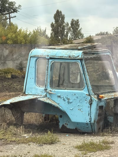 The image shows the cab of an old, light blue truck, abandoned and weathered. The paint is heavily chipped, revealing rust underneath, and the windows are dirty, obscuring the interior. The cab appears to be sitting on the ground, with the rest of the truck missing, suggesting it's been dismantled or is in the process of decaying.

The setting is an outdoor, rural or semi-rural environment, likely in Samhorodok, Ukraine. In the background, there are trees and vegetation, some of which are dry and overgrown, further indicating abandonment. Overcast skies and a hazy atmosphere suggest it might be a cloudy day, possibly damp given the weathered appearance of the truck. There are power lines visible in the distance, indicating proximity to a populated area or infrastructure, but the immediate surroundings are neglected. The overall mood is one of decay, abandonment, and the passage of time. No people are visible in the image, and no text is discernable.