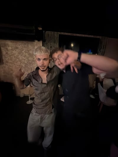 The image depicts two individuals in a dimly lit indoor setting, possibly a bar or lounge. The location cannot be definitively identified as Slivnitsa, Bulgaria, based on visual evidence within the image.
On the left, a male with short, bleached blonde hair and a visible septum piercing is captured with moderate clarity. He has a light mustache and goatee. He wears a light gray or tan short-sleeved collared shirt, unbuttoned at the top, over a dark undershirt or chest. He also wears light-colored trousers. His right arm is raised with an open hand gesturing towards the camera, while his left arm is bent, also in a gesturing posture. He looks directly at the camera with a relaxed expression.
To his right, a second male figure is heavily blurred due to motion. He appears to wear glasses and a dark-colored shirt, with a dark wristband or watch visible on his left wrist. His left arm is raised and bent, with the hand in a partial claw shape, while his right arm is also raised, partially obscuring his face. This individual is positioned slightly behind and to the right of the first.
The background on the left features a textured, light brown or beige wall. A white, vertical stand-like object is visible near the floor behind the left individual. The background on the right includes vertical striped elements, possibly wall decor or blinds. Further back, a dark area reveals faint light sources and a distinct blue light, possibly from a screen or an external light fixture. Partially visible on the far right are white and light-colored chairs with wooden legs, indicating a seating area. The overall lighting is low, with the primary illumination appearing to originate from the front, slightly to the left of the scene.