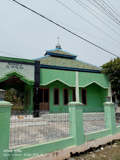 The image shows a small mosque in Kecamatan Gabuswetan, Indonesia. The mosque is a single-story building with a green exterior and a tiled roof. It has a small dome on top and a sign above the entrance that reads "Musholla Al-Ikhlas". The mosque is surrounded by a chain-link fence and a concrete wall. There are some trees and power lines in the background. The scene is taken from the perspective of someone standing in front of the mosque. There is a small, yellow object in the yard, possibly a piece of trash or a small animal. There is also a white sign in the lower right corner of the image, with the words "VIVO Y19s" and "Akhtar & Arvan 10 Apr 2025, 10:52".