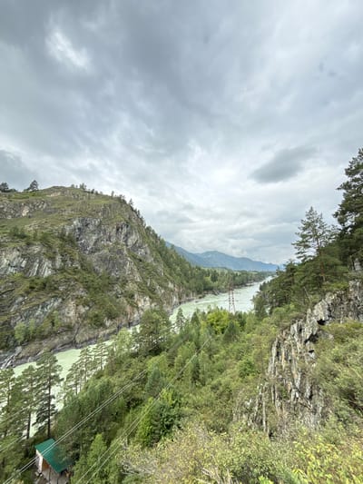 The image shows a wide river flowing through a valley surrounded by steep, rocky mountains covered in lush green vegetation and pine trees. The sky is overcast with heavy gray clouds. In the foreground, a small wooden building with a green roof is visible on the left side, nestled amongst trees and a power line runs diagonally across the scene. In the middle ground, the river curves to the right, with power lines crossing it. Further in the distance, mountains form a hazy backdrop. The scene appears to be a natural landscape in a mountainous region, possibly in or near Chemal, Russia, given the description of the location. There are no people visible in the image.