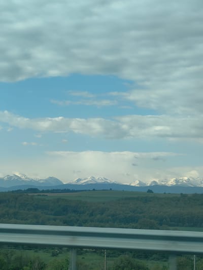 The image shows a landscape view near Vakarel, Bulgaria. The foreground features a highway guardrail, indicating the photo was likely taken from a moving vehicle. Beyond the guardrail, there's green vegetation, possibly fields or a low-lying forest. In the midground, a line of wooded hills extends across the frame. The background showcases a range of snow-capped mountains under a sky filled with a mix of white and grey clouds, with patches of blue sky visible. The overall impression is of a rural landscape seen from a highway.