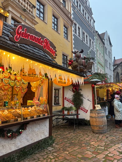 The image displays an outdoor Christmas market scene in Meissen, Germany, with a cobblestone ground under an overcast sky.
In the foreground and midground, a market stall prominently features the illuminated sign "Gebrannte Nüsse" (roasted nuts) in red script with a white border on a dark brown backdrop. The stall's awning is adorned with white, icicle-shaped cutouts and string lights. Its display shelves are laden with various roasted nuts and candies. Gingerbread hearts, some labeled "Liebesäpfel" and others with German messages, hang from the interior. A festive garland with red spherical ornaments drapes the lower front of the stall. A plush reindeer figure, dressed in a plaid scarf and pulling a small sleigh containing red ornaments and greenery, is mounted on the roof above the right side of the "Gebrannte Nüsse" stall.
To the right, a black folding table with benches is situated on the cobblestones. Adjacent is a second, partially visible market stall, displaying a sign reading "Winzer...sion - Wein," decorated with evergreen branches and red bells. A weathered wooden barrel stands directly in front of this stall.
One female individual is visible in the midground to the right, wearing a light-colored, possibly mint green, long puffer coat and a pink knit beanie. Her head is turned slightly to the left. Blurred figures are discernible in the deeper background.
The background features a row of multi-story buildings: a yellow-facade building directly behind the "Gebrannte Nüsse" stall, followed by a gray building, then lighter green and cream-colored structures, all with visible windows. A dark, gothic-style architectural structure with a pointed roof is partially visible in the far background on the right.