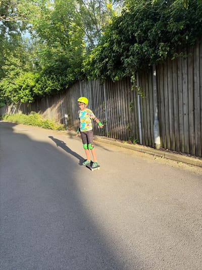 A young person wearing a yellow helmet and protective pads is skateboarding on an asphalt path. The path is lined by a wooden fence and dense green foliage. The sun is casting a long shadow from the skateboarder. The scene appears to be in a park or recreational area.
