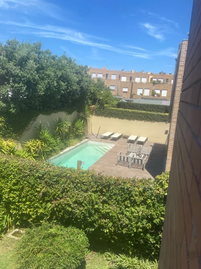 The image shows a courtyard with a swimming pool and loungers. The scene is set in a sunny location, likely outdoors, given the bright blue sky and strong shadows. The architecture suggests a residential or hotel setting, possibly in Matosinhos, Portugal, as indicated by the context.
The main subjects are the swimming pool, which has light blue water, and a wooden deck area surrounding it. Several lounge chairs are arranged on the deck, some with cushions, suggesting a place for relaxation. Lush green vegetation, including trees and bushes, frames the courtyard, creating a private and serene atmosphere. In the background, a multi-story building with white-trimmed windows is visible.
The time of day appears to be late morning or early afternoon, judging by the intensity of the sunlight and the length of the shadows. The weather is clear and sunny, with a few wispy clouds in the sky. There is no visible text in the image. The overall impression is one of a peaceful and inviting outdoor space, perfect for enjoying a warm day.