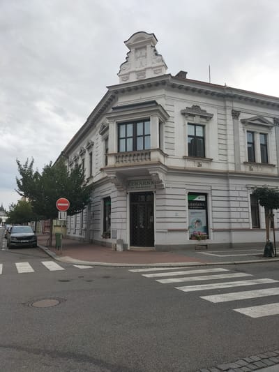 The image captures a street corner in Pardubice, Czechia, featuring a white, ornate building that houses a pharmacy. The building has classical architectural elements like decorative pediments, columns, and recessed window frames. A sign above the entrance reads "LÉKÁRNA ve Smilověce." A pedestrian crossing is visible in the foreground, marked by white stripes on asphalt. To the left of the building, a black car is parked on the street, and a red and white circular traffic sign is posted near a tree. The sky is overcast, suggesting a cloudy day. The scene is devoid of any visible people or significant activity.