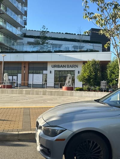 In West Vancouver, Canada, on a clear, sunny day, a silver BMW 3 Series sedan is parked in front of a modern building complex. The building features a multi-story residential structure with glass balconies and a ground-level commercial space. The commercial facade displays the "URBAN BARN" signage, indicating a retail establishment. In front of the building, there are two abstract sculptures on red bases, a trash receptacle, and several large windows, some of which are covered with frosted film or display advertisements for "R-MEDY YOUR FAVOURITE COSMETIC CLINIC." A sidewalk with brick paving separates the parking area from the building's entrance. The environment is well-maintained with landscaping, including trees and shrubs. The sky is a clear, bright blue, suggesting daytime.