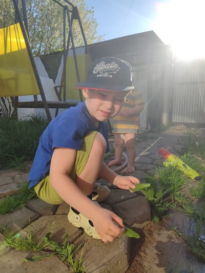 A young boy in a blue shirt, green shorts, and a black "Skate" hat sits on a brick-paved area outdoors, holding green leaves in his hands. He wears light-colored Croc-style shoes. Behind him, another child in a striped yellow and gray shirt and diaper stands near a garden hose. A yellow and red water gun lies on the ground among the grass. A swing set with yellow fabric is visible in the background, along with a fence and trees. The sunlight is bright, casting shadows across the scene. Location is stated as Malaya Purga, Russia.