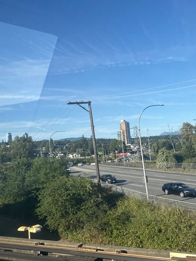 The image captures a view of a highway with traffic, bordered by dense green foliage and trees. In the distance, several buildings, including a prominent tall, modern skyscraper, are visible against a clear blue sky with wispy clouds. A utility pole with wires and insulators stands in the foreground, with streetlights lining the highway. The scene suggests an urban or suburban environment, likely New Westminster, Canada, given the architectural style and natural landscape. There are no individuals clearly visible in the frame. The perspective appears to be from an elevated vantage point, possibly a bridge or a higher level of a building.