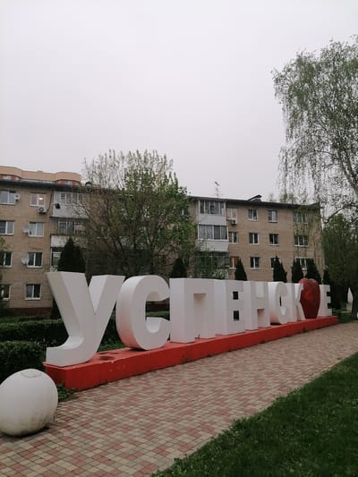 The image depicts a large, freestanding sign in the city of Uspenskoye, Russia. The sign spells out "УСПЕНСКОЕ" in large, white Cyrillic letters, mounted on a red base. A red apple figure replaces the letter “O”. The sign sits on a paved area composed of red and brown patterned bricks. Behind the sign, there's a multi-story brick building with several windows and balconies. Trees and bushes surround the sign and the building. The sky is overcast. A large, white, spherical object sits on the pavement next to the sign. A grassy area is visible to the right of the paved area.