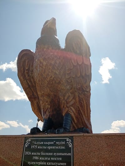 The image shows a large, wooden sculpture of an eagle, likely a monument, against a bright blue sky with fluffy white clouds. The sun is shining intensely, creating lens flare and casting shadows, suggesting it is daytime. The eagle sculpture is positioned centrally, viewed from below, with its wings spread wide and its head turned slightly upwards. The texture of the sculpture appears to be detailed, mimicking feathers and the structure of an eagle's body.

In the foreground, a granite base supports the sculpture, and on this base, a dark plaque with white lettering is visible. The plaque contains text in a non-Latin script, likely Kazakh. The text on the plaque appears to be a dedication or informational inscription related to the sculpture. Part of the text reads, "“Алтын қыран” мүсіні 1975 жылы орнатылды." which translates to "Golden Eagle sculpture installed in 1975." Other lines mention "2026 жылы Балқаш ауданының 1986 жылғы мектеп түлектерінің қатысуымен" which translates to "with the participation of the 1986 school graduates of Balkhash district in 2026."

The overall scene is grand and patriotic, emphasizing the significance of the "Golden Eagle" monument. The bright, sunny weather and clear sky contribute to a positive and uplifting atmosphere. The location context provided is Baqbaqty, Kazakhstan.