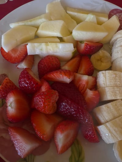 A close-up overhead shot shows a plate of sliced fruit, including apples, bananas, and strawberries. The apples are cut into rectangular segments, the bananas into round slices, and the strawberries mostly halved or quartered. There are no people visible. The plate appears to be ceramic and rests on a red surface. The location is not indicated and not discernable from the image.