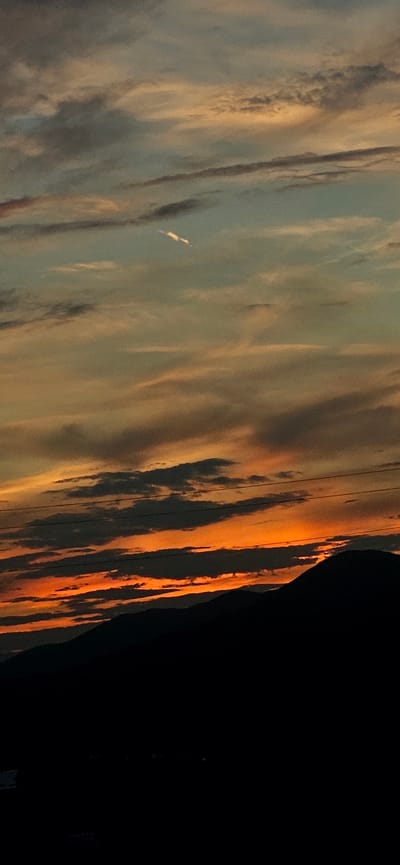 The image depicts a dramatic sunset over the city of Momin Prohod, Bulgaria. The sky is filled with vibrant hues of orange, yellow, and red, painted across a canvas of soft, wispy clouds. Silhouetted against the colorful sky is a range of dark, undulating mountains, their peaks softened by the twilight. In the foreground, the landscape is plunged into darkness, obscuring any finer details of the city below. Two thin, dark lines, likely power lines, stretch horizontally across the middle of the frame, adding a man-made element to the natural beauty of the scene. The overall impression is one of natural splendor and serene tranquility.