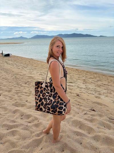 A medium-shot, full-body image shows a Caucasian woman with shoulder-length blonde hair, wearing a leopard-print one-piece swimsuit and carrying a large leopard-print tote bag with rope handles. She is standing on a sandy beach, facing the viewer and smiling. The ocean is in the background, with a distant view of islands and a cloudy sky. A small propeller-driven device, possibly a jet ski or an electric surfboard, is visible on the sand to the left of the woman. The lighting suggests it is daytime, possibly late afternoon. The location appears to be a beach in Ko Samui District, Thailand, given the presence of tropical islands and the style of the device on the beach.