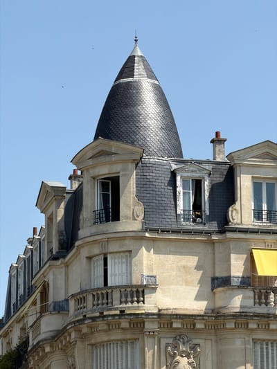 The image shows a close-up, low-angle view of the mansard roof and upper floors of a stone building with a conical grey shingle turret in Neuilly-sur-Seine, France. The sky is clear blue with two small birds visible in flight. The building features multiple windows with various styles of shutters and balconies, including a rounded balcony with ornate balusters on the right side of the frame. A yellow awning is partially visible on the right. Chimneys with red caps protrude from the roofline. The architectural style suggests a turn-of-the-century Parisian apartment building.