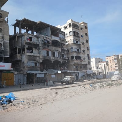 The image captures a street scene in Jabalia, Palestinian Territory, under a clear blue sky with sparse white clouds. The foreground features an unpaved, rubble-strewn road heavily littered with debris, rocks, and refuse, including a prominent pile of blue plastic bags on the left.
The left side of the street is lined with multi-story buildings displaying significant structural damage. A central structure shows severe destruction to its upper floors, including exposed rebar, collapsed sections, and missing windows. Laundry is visible hanging from balconies and makeshift lines on various levels. At its ground level, several makeshift stalls or enclosures, covered by tarpaulins and canvas, are present, with several indistinct figures visible within these shaded areas. A small individual stands near a yellow-green door or gate on the far left.
To the right, a taller, lighter-colored multi-story building, approximately seven to eight floors high, exhibits signs of facade damage, including discoloration and some broken windows, but appears more structurally sound than its immediate neighbor. Further down the street, additional multi-story buildings are visible in the background, some appearing intact, others with signs of wear.
A light-colored 4x4 utility vehicle is positioned on the right side of the road. Further in the mid-ground, a damaged white sedan is partially visible.