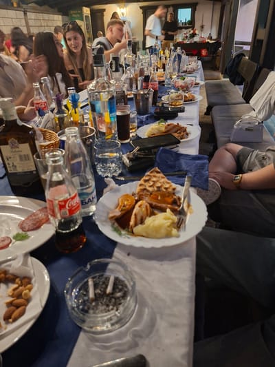 The image depicts an indoor social gathering, likely a dinner or party, at a long table set for numerous guests. The perspective is slightly elevated, looking down the length of the table which is adorned with a white tablecloth and a blue runner.

Numerous people are seated around the table, engaged in conversation and enjoying food and drinks. In the foreground-left, a person wearing a grey shirt and dark pants is partially visible, sporting a gold digital watch on their wrist. Further down the table, a man with short hair and a striped shirt is seen, along with a woman with long dark hair who is looking towards the camera with a slight smile, suggesting a convivial atmosphere. Other blurred figures indicate a bustling event.

The table is laden with various dishes, including plates of grilled meat, mashed potatoes, a waffle-like pastry, salads, nuts, and sliced sausage. A prominent clear glass ashtray containing cigarette butts and ash is visible on the right side of the table. A wide selection of beverages is present, including small glass bottles of Coca-Cola in an ice bucket, clear bottles with Cyrillic text "БАН" (likely water), and a bottle of Ballantine's whiskey. Numerous clear drinking glasses and other bottles containing spirits or juices are also scattered across the table. A blue plastic bag or container is also on the table.

In the background, upholstered seating and a window or doorway are visible. On a distant table to the left, several gifts wrapped in red and white are present. The lighting suggests an evening event. A sign in the background features Cyrillic text, reading "ДУХЪТ НА ПРАГА" (The Spirit of Prague), and other Cyrillic labels are visible on various bottles, consistent with the location context of Ruska Bela, Bulgaria.