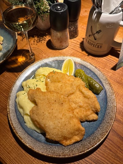 This image captures a close-up, high-angle view of a meal served on a wooden table, likely in a restaurant or pub setting. The primary focus is a blue-grey ceramic plate holding what appears to be two large, golden-brown breaded cutlets, similar to schnitzel. These cutlets are crispy in texture and rest on a bed of creamy mashed potatoes, which are lightly garnished with chopped green herbs, possibly parsley.
Accompanying the main dish on the plate are two small green gherkin pickles and a bright yellow lemon wedge, positioned near the top left of the plate. The plate itself has a distinctive, rustic-looking rim with an uneven, textured finish in shades of brown and grey.
To the left of the plate, several other items are visible on the warm-toned wooden table. In the upper left quadrant, a white ceramic mug or pitcher with a handle is seen. It features a dark graphic logo depicting a crossed fork and knife, below which the words "HOSTINEC" and "NA KOPEČKU" are printed in dark capital letters. The mug has some condensation or reflections on its surface.
Below the mug, two cylindrical spice grinders, likely for salt and pepper, are placed. They have black tops and clear bodies, allowing their contents (coarse salt and peppercorns) to be seen. A small decorative plant with green foliage and white accents is partially visible just to the left of the grinders.
Further down and to the left, a stemmed wine glass, filled with a light-colored liquid (suggesting white wine), is prominent. Another plate, partially obscured, is visible below the wine glass.
The lighting in the scene is warm, casting some soft shadows, particularly from the objects on the left side of the table, suggesting an indoor setting, possibly during the evening. The wooden table surface has a visible grain, adding to the rustic aesthetic of the scene. The overall impression is one of a prepared meal waiting to be enjoyed.