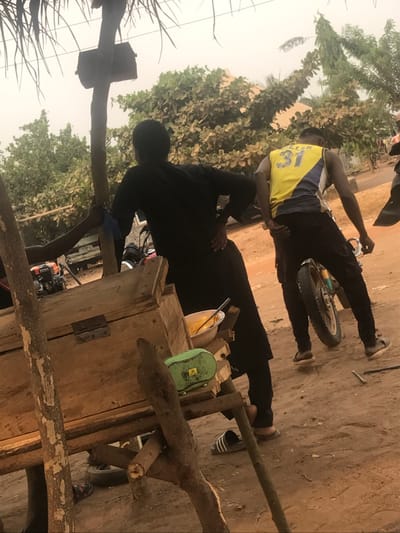 Two men are outside on a sandy surface. One man, wearing a yellow jersey with "31" on the back and black pants, stands near a bicycle wheel. The other, dressed in black, appears to be working near a wooden structure, possibly a cart. A green object and a yellow container sit on top of the wooden structure. The scene includes trees and foliage in the background.