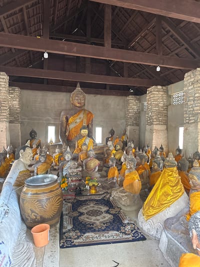 This image depicts the interior of a temple in Baan Harn, Thailand, housing numerous Buddha statues. The statues vary in size and material, with many adorned in saffron robes. A large, central Buddha statue is prominent, surrounded by smaller ones arranged in rows and clusters. In the foreground, a large ceramic jar with a painted dragon motif sits next to an orange bucket. A patterned rug is laid out on the floor in front of some of the statues. The architecture features exposed wooden beams supporting a tiled roof and stone pillars. Sunlight streams in through rectangular openings in the walls, illuminating the scene.