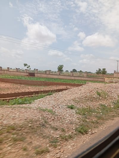 The image captures an outdoor scene, likely a construction site or agricultural land, under a partially cloudy sky. In the foreground, a textured surface of small, light-colored stones dominates the right side, with patches of green vegetation interspersed. To the left and center, there are demarcated plots of land. Some plots are bare earth, while others contain rows of green plants, possibly crops, and are bordered by brick walls. A more substantial wall with a decorative white railing runs horizontally across the mid-ground, separating this area from a distant background. Beyond this wall, more trees and vegetation are visible, along with a utility pole on the far right. The overall impression is of an undeveloped or developing area with signs of agricultural activity. The scene is devoid of any people. The location is specified as Wuro Dole, Nigeria, suggesting an arid or semi-arid environment with efforts towards land development and cultivation.