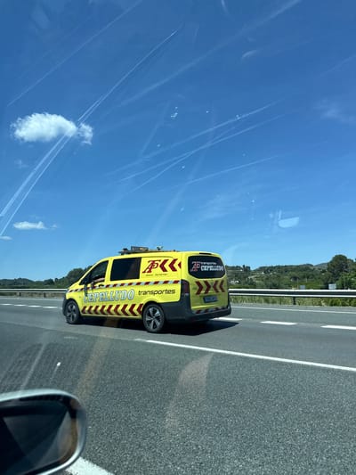 A yellow van with the name "CEPELLUDO" and "transportes" written on it is driving on a road in Rotglà i Corberà, Spain. The vehicle has a license plate of "1064 MVS". The road is marked with white lines. The sky is blue with contrails and a single cloud. The landscape includes green hills in the background. The image appears to be taken from inside another vehicle, with the side mirror partially visible.