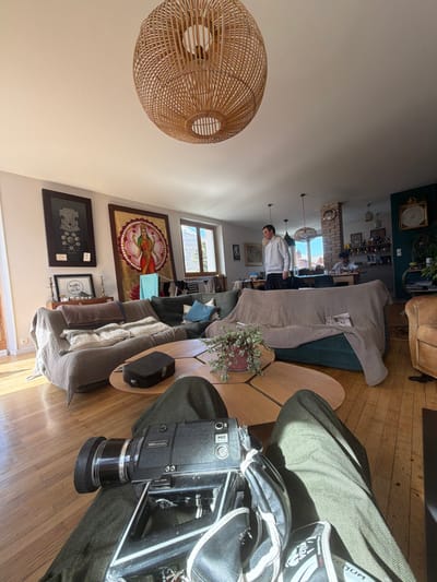 This image depicts a residential living room and an adjacent kitchen in Briançon, France, viewed from a first-person perspective. In the immediate foreground, a person's legs, dressed in dark olive-green trousers, are visible. Resting on their lap is a black "Mirage M8" Super 8 film camera with a large lens, accompanied by a pair of white and black "Venum" branded sports gloves.

The mid-ground features a multi-segment coffee table constructed from light-toned wood, on which a small black hard case and a potted plant with green leaves are placed. Two large sofas, one light brown with a textured throw and another dark green with a similar covering, are arranged around the coffee table.

In the background, a man with dark hair, wearing a light-colored hooded sweatshirt and dark trousers, stands in an upright posture between the living room and kitchen areas, looking towards the right. Further into the kitchen, a second individual is seated at a table, wearing a light-colored top. The living room walls are light-colored and display two framed artworks: a monochromatic print with circular patterns and a large, colorful painting depicting a multi-armed deity in a lotus. A wooden console table with two metallic candle holders is situated beneath these artworks. A large, woven wicker pendant lamp hangs from the ceiling in the upper-center. Bright natural sunlight streams through two windows on the left wall, illuminating the light wooden parquet flooring and revealing distant snow-capped mountains. The kitchen area features dark green walls, a brick pillar, open shelving with various items, and additional pendant lighting.