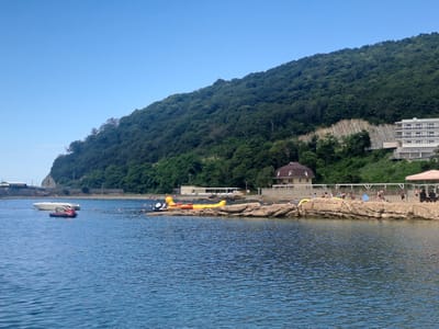 The image shows a coastal scene in Dzhubga, Russia. The foreground is dominated by calm blue water. In the middle ground, a rocky shoreline extends into the water. Several inflatable water toys, including a yellow banana boat and a red jet ski, are visible on the water and near the shore. A white boat is anchored in the distance.
The background features a lush green hillside that slopes down towards the water. A white multi-story building, identified by the sign "ПАРК-ОТЕЛЬ" (Park-Hotel), is situated on the hillside. Several people are visible on the beach and near the water, engaged in recreational activities. The sky is clear and blue.