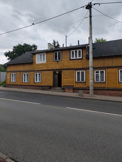 The image depicts a two-story wooden building with yellow siding, located at 24 Słowackiego Street in Zgierz, Poland. The building has a dark-colored, sloped roof and features numerous white-framed windows on both levels. An open doorway is visible on the right side of the ground floor, leading to steps. A concrete utility pole with wires and a street lamp stands in front of the building, positioned slightly to the right of center. The foreground shows an asphalt road with a white line marking the edge of the traffic lane, adjacent to a paved sidewalk. The sky is overcast and cloudy. A dark bird is perched on a wire in the upper left quadrant of the image.