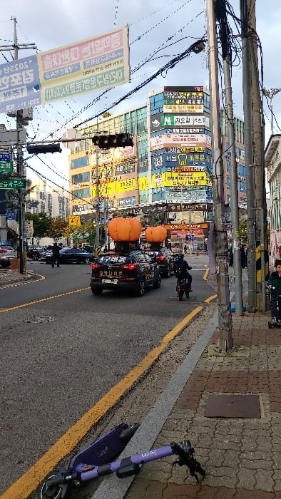 The image depicts an urban street scene in Gimpo-si, South Korea, under an overcast sky. In the foreground on the right, a purple Lime electric scooter lies horizontally on a patterned sidewalk next to a yellow curb.

In the midground, a street curves slightly to the left. Two black SUVs, prominently featuring large orange, pumpkin-shaped inflatable decorations on their roofs, are observed in a line. The lead SUV displays Korean text including "구래동" and "김포나이트" on its side. Following these vehicles is a person on a black motorcycle. A horizontal traffic light fixture with multiple lenses is visible above the street. On the far left, several cars are parked alongside the curb, and a person dressed in dark clothing stands near them.

On the right sidewalk, partially obscured by a utility pole, a young child wearing a green jacket is seen riding a smaller scooter.

The background is dominated by multi-story commercial buildings heavily adorned with numerous vertical and horizontal signs in Korean, exhibiting a wide array of colors and advertisements for various businesses. Overhead, numerous electrical and communication wires crisscross the street, suspended from utility poles. A large banner with Korean text is also strung across the street. Multiple utility poles line the right side of the street. The road surface is asphalt with yellow painted lines.