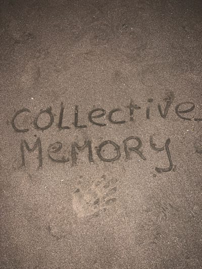 This image presents a top-down, close-up view of a light brown, granular sandy surface in Kerobokan Kelod, Indonesia. The words "COLLECTIVE MEMORY" are inscribed into the sand across two lines, with "COLLECTIVE" centered above "MEMORY." The letters are capitalized and formed by depressions in the sand. Below and to the left of the word "MEMORY," a distinct impression of a human left hand is visible in the sand, showing outlines of the palm and fingers. No individuals or external objects are present in the frame. The sand appears uniformly illuminated, highlighting its texture and the carved inscriptions and print.