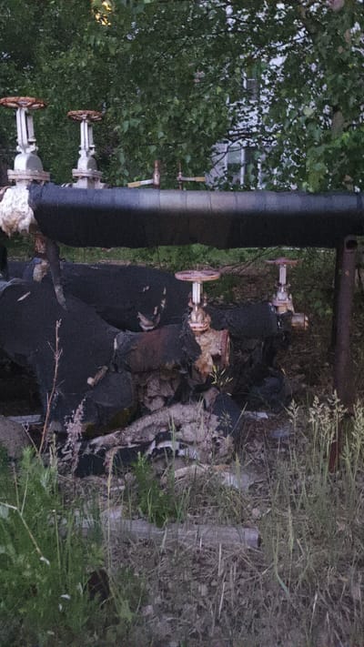 The image depicts an outdoor scene in Kogalym, Russia, featuring industrial piping and valves amidst overgrown vegetation. A large, black insulated pipe runs horizontally across the upper portion of the frame, supported by metal structures. Below this, a complex network of smaller, black-covered pipes and rusted valves is visible. Some of these lower pipes appear damaged or degraded, with insulation peeling away and exposed metal. The ground is covered in a mix of dirt, debris, and tall, wild grass, indicating a neglected or abandoned industrial area. Trees with dense green foliage form a backdrop, partially obscuring a building in the distance. The overall impression is one of disuse and the encroachment of nature on industrial infrastructure.