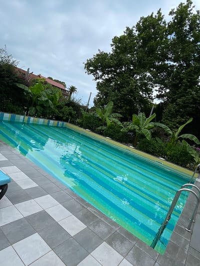 The image shows an outdoor swimming pool with a striped pattern in shades of green and blue. The pool is surrounded by a tiled patio with a checkerboard pattern of gray and white squares. Several banana trees and other lush green vegetation are visible behind the pool. In the background, a building with a red tiled roof can be seen. The sky is overcast. The scene appears to be a resort or a private residence in Abaata, Abkhazia. There are no people visible in the image, nor any actions or interactions taking place.