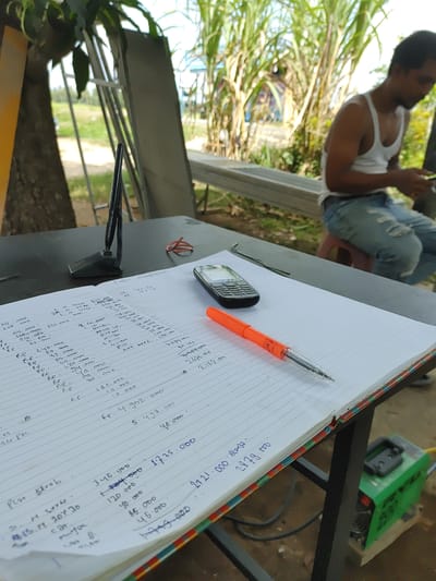 The image depicts a close-up, slightly tilted view of a table in an outdoor setting, likely in Indonesia given the context. The main subjects are a person sitting in the background and various objects on the table in the foreground.
The person in the background is a man wearing a white tank top and ripped blue jeans. He is partially obscured by the foreground elements and appears to be looking down at something, possibly his phone. The background scene suggests a casual, possibly work-related environment, with lush green foliage and what appears to be construction or industrial materials like a green welding machine and metal frames. The lighting suggests it is daytime.
The foreground is dominated by an open notebook filled with handwritten notes and calculations. A bright orange pen rests on the page, indicating that someone was recently writing or is about to write. A black mobile phone is also placed on the notebook. The notebook's pages are lined and contain numerous entries with numbers, some of which appear to be currency amounts, and brief descriptions. The visible text includes phrases like "PINTU HANDERSONS," "Cat, dowr," "Silikon," "Plat Streb," and various numerical figures that could represent costs or quantities. The edge of the notebook shows colorful striped binding.
To the left of the notebook, a black tool with a textured grip is positioned, possibly a clamp or a part of some equipment. A small, red circular object, possibly a rubber band or a small lid, is also visible on the table.
The overall impression is one of a work in progress, a moment captured while someone is engaged in calculations or record-keeping in a relaxed, outdoor environment. The combination of the person, the notebook, and the tools suggests a task related to construction, repair, or inventory.