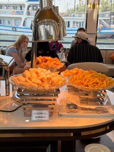This image depicts a buffet setup inside what appears to be a boat or cruise ship, offering a view of the outside environment. The photograph is oriented vertically, rotated 90 degrees clockwise.

In the foreground and mid-ground, a selection of fried foods is displayed in multiple clear, decorative glass dishes. These include a large quantity of golden-brown french fries, a bowl of bright orange, puffed snacks, and two bowls filled with golden-brown, cylindrical items. Adjacent to these, a small white sign clearly labels them as "SEAFOOD SPRING ROLLS" and "Nem hải sản chiên" (Vietnamese for "Fried seafood spring rolls"). Metal serving tongs are neatly arranged on a rack next to the food, ready for use. The food is kept warm in polished metal chafing dishes, one of which features a yellow warning symbol for heat.

In the background, several individuals are present. A man wearing a striped shirt, a white baseball cap, and sunglasses pushed up is seated. To the lower left, a woman with blonde hair, wearing a patterned dress, is also seated, looking towards the right. The setting appears to be a comfortable dining area with large windows.

Beyond the interior, a bright, sunny daytime scene unfolds. The windows offer a clear view of a body of water, with other boats visible, including a prominent white vessel with blue trim and Vietnamese text and numbers (partially legible) on its side. Palm trees and distant land can also be seen, indicating a scenic coastal or island location. The scene suggests a relaxed dining experience on a cruise or tour boat in Tuan Chau Ward, Vietnam.