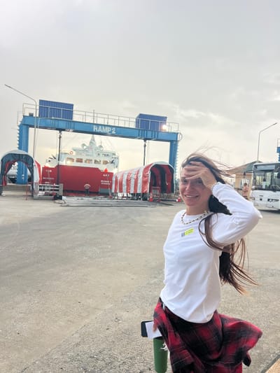 In Don Sak, Thailand, a woman stands on a concrete surface, her face illuminated by soft daylight. She wears a white long-sleeved shirt and a red and black plaid skirt. Her hair is windswept. Her right hand is raised, touching her forehead. Behind her, a red ship with white accents is docked. A blue gantry crane with "RAMP 2" labeled on it is positioned over the ship. Red and white striped tents are set up near the ship. A white bus is visible to the right of the frame, and another person, possibly a man, is seen in the background near a building. The sky is overcast.