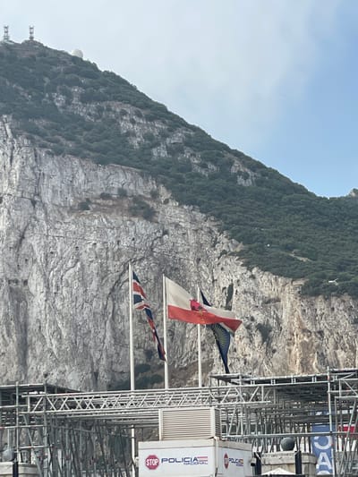 The image shows a rocky mountainside with sparse vegetation, under a clear sky. Two flags are prominently displayed in the foreground, fluttering from poles. The flag on the left appears to be the Union Jack, while the flag on the right is white with a red emblem in the center and a dark blue, possibly black, streamer hanging below. In the lower part of the image, there is scaffolding and structures that suggest ongoing construction or event setup. A white container unit with "STOP POLICÍA NACIONAL" text is visible. The context of La Línea de la Concepción, Spain, suggests this view is likely of the Rock of Gibraltar, which is directly across the border from La Línea.