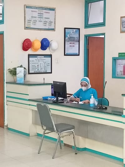 The image shows the interior of a building in Kecamatan Margorejo, Indonesia (latitude: -6.76, longitude: 111.015). A woman in a blue shirt and white head covering is seated at a desk writing on a piece of paper. There is a computer monitor and a hand sanitizer bottle on the desk. Behind her is a closed door, and to the left, a counter and a chair. The walls are white, and there are four hard hats, a framed chart, and a framed poster on them. There is a potted plant in the corner and a small framed bulletin board on the right wall. The floor is tiled.