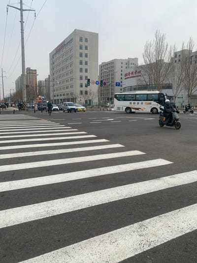A slightly tilted outdoor shot captures a street scene with a crosswalk. White stripes contrast sharply against the gray asphalt. A white bus and several motorcycles are on the road, lining up at a traffic light. Buildings in varying heights form the backdrop under a slightly overcast sky. The composition focuses on the repetitive pattern of the crosswalk, adding a graphic element to the urban environment.