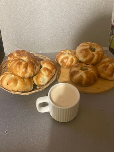 A top-down view shows a breakfast spread on a grey countertop. In the center of the frame, a white mug with a ribbed texture is filled with a light-colored beverage, possibly milk or coffee with milk. To the left of the mug, a wooden cutting board holds five golden-brown baked goods. These pastries appear to be filled, with visible openings revealing a hint of their contents. Three more similar pastries are arranged in a woven basket to the lower left of the cutting board. The pastries have a glossy sheen, suggesting they may have been brushed with egg wash or butter. The scene is likely indoors, given the countertop and the presence of a mug. The lighting is overhead, creating soft shadows. The overall impression is one of a simple, homemade breakfast. No people are visible in the image. No text is present in the image.