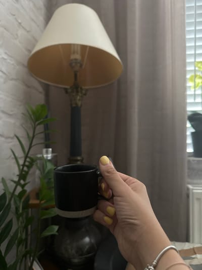 The image depicts a close-up view of a person's hand holding a black mug. The hand has bright yellow nail polish. In the background, a lamp with a cream-colored shade is visible, along with a curtain and a white brick wall. A green plant is also present in the scene. The setting appears to be indoors, possibly a home environment. There is no information to indicate the location in the city of Macclesfield, United Kingdom.