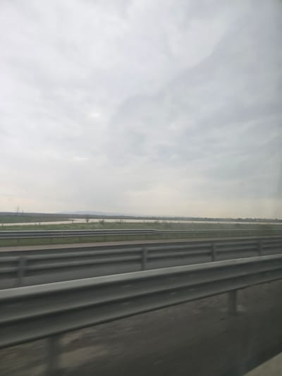 The image displays a highway landscape under an overcast sky, located in the vicinity of Opalchenets, Bulgaria. The foreground features a multi-lane asphalt highway, characterized by several parallel rows of grey metallic crash barriers extending horizontally across the lower portion of the frame. The perspective indicates the photograph was taken from a moving vehicle, looking slightly forward and to the right. Beyond the highway, a narrow strip of green grass precedes a wide, placid body of water, which reflects the pale, diffused light of the sky. Sparse, bare deciduous trees are visible along the distant bank of the water. On the horizon, a low, indistinct line of hills or mountains is visible beneath a uniformly light grey, cloud-covered sky. No individuals, explicit actions, or specific interactions are depicted in the scene.