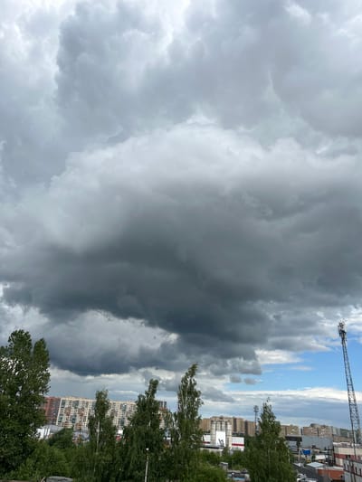 This is a vertical, eye-level shot of a cloudy sky over a residential area in Bugry, Russia. The dominant feature is a dramatic expanse of dark, heavy storm clouds that take up the majority of the frame, obscuring much of the sky. To the right, there are patches of blue sky visible, with lighter, more scattered clouds.
Below the clouds, the scene transitions to a cityscape. Buildings, primarily multi-story apartment blocks, are visible in the mid-ground and background. These buildings appear to be residential, with many windows and balconies. The architecture suggests a modern urban or suburban setting.
In the foreground and along the right side of the image, lush green trees, possibly birch trees, line the edge of the view, their leaves rustling. The trees are dense and add a natural element to the otherwise urban landscape.
Visible in the upper right of the frame is a tall, lattice-like metal structure, possibly a communication tower or a construction crane. Another similar, but smaller, tower is visible further into the scene.
The weather appears to be stormy or overcast, with the dark clouds suggesting the possibility of rain. The lighting is diffused due to the cloud cover, and there are no harsh shadows. The overall mood is somber and perhaps a little dramatic, given the imposing clouds. The time of day is difficult to determine precisely due to the lack of direct sunlight, but it appears to be daytime. No people are visible in the frame. There is no visible text.