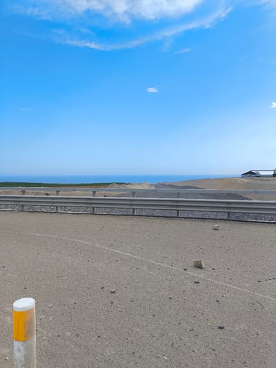 The image depicts a roadside scene in Huanchaco, Peru, characterized by a clear blue sky with scattered clouds and a distant view of the ocean. A grey metal guardrail runs horizontally across the midground, separating the foreground from the backdrop. Behind the guardrail is a gravelly embankment. The foreground is a grey asphalt surface with scattered small rocks and a short, white and yellow marker post on the left. A building or structure is visible in the far background. No people are present in the image.