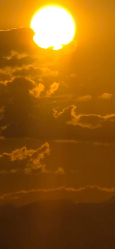 This image depicts a vivid sunset over Basbeshik, Kyrgyzstan. The sun, a brilliant white orb with a bright halo, dominates the upper portion of the frame, partially obscured by wispy, golden clouds. The sky transitions from a luminous yellow near the sun to a deeper orange and then to a dark, hazy brown at the bottom. The lower third of the image is a silhouetted landscape, likely mountains or hills, appearing as indistinct dark shapes against the glowing sky. There are no people or specific man-made objects visible. The scene is characterized by its warm, intense colors and the dramatic interplay of light and shadow from the setting sun.