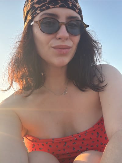 The media file shows a close-up, upside-down selfie of a person outdoors. The subject has dark hair, wears sunglasses with round, tortoiseshell frames, and a leopard-print headscarf. They are also wearing a red bikini with a black dot pattern. A thin silver necklace with a constellation pendant is visible on their neck. The scene appears to be a sunny day, with bright light casting a glow on the subject's hair and skin. The background consists of a clear blue sky, suggesting an outdoor setting like a beach or a boat. The overall mood conveyed is casual and relaxed, indicative of a vacation or leisure activity. The bright sunlight suggests it is daytime. The location context of Salou, Spain, further supports the idea of a sunny, coastal destination.