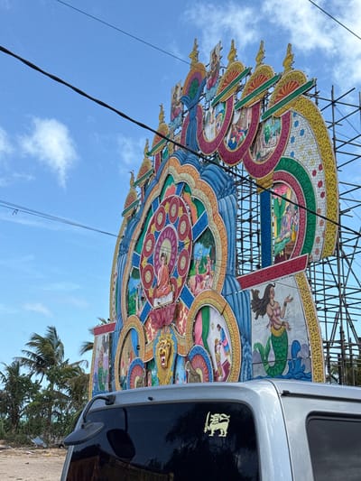 This is a medium shot, eye-level, outdoor photograph taken in Habaraduwa, Sri Lanka. The main subject is a large, ornate, colorful archway made of painted panels supported by scaffolding. The archway depicts various scenes and figures, including a Buddha figure at the center, a lion, and a mermaid. The archway is situated outdoors, with a blue sky and scattered white clouds visible above. Palm trees and vegetation are seen in the background, along with debris on the ground, suggesting a possibly undeveloped or coastal area. A portion of a silver vehicle is in the foreground, obscuring the lower part of the archway and the ground. The vehicle has a tinted rear window with a yellow sticker of a lion holding a sword. Power lines are visible across the sky and in front of the archway. The image appears to be taken during daylight.