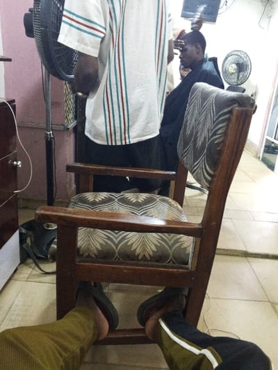 The scene is an interior view of what appears to be a barbershop in Oko Erin, Nigeria, observed from a low, seated perspective. In the foreground, the lower legs and feet of the photographer are visible, wearing olive green trousers on the left and dark trousers with a white stripe on the right, both with dark slip-on shoes. Immediately in front of the photographer is a wooden chair with dark brown arms and legs, featuring a patterned seat and back cushion in beige and dark brown leaf and scroll motifs.
Beyond this chair, a male barber stands with his back mostly towards the camera, wearing a white short-sleeved shirt with vertical red and teal stripes, over dark trousers. His right hand, adorned with a ring, is visible near his waist. To the barber's left, a large black standing fan is positioned. To the barber's right, a male client is seated in a dark chair, covered by a black barber's cape, and is in the process of receiving a haircut. A barber's tool, possibly clippers, is held near the client's head by an unseen hand, likely the barber's. Another black standing fan is visible behind the client.
The walls are painted a light purple-pink color. The floor is tiled with light-colored square tiles. In the background, a blurry wall-mounted screen or monitor is visible, along with general indoor lighting. A dark bag rests on the floor next to a dark wooden cabinet on the left side of the frame. White electrical cords are visible on the floor and against the cabinet.