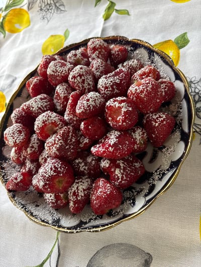 A close-up overhead shot shows a bowl filled with strawberries dusted with powdered sugar. The strawberries are a vibrant red and are piled high in a decorative bowl with a gold trim and a dark blue and white patterned rim. The bowl rests on a tablecloth with a repeating pattern of yellow lemons and green leaves, set against a textured, neutral background. The lighting suggests daytime, possibly indoor lighting as there are no strong shadows. The overall impression is one of a simple, appealing dessert or snack. There is no visible text in the image. The location context is provided as Permyak, Russia, though there are no specific visual cues within the image to confirm this location.